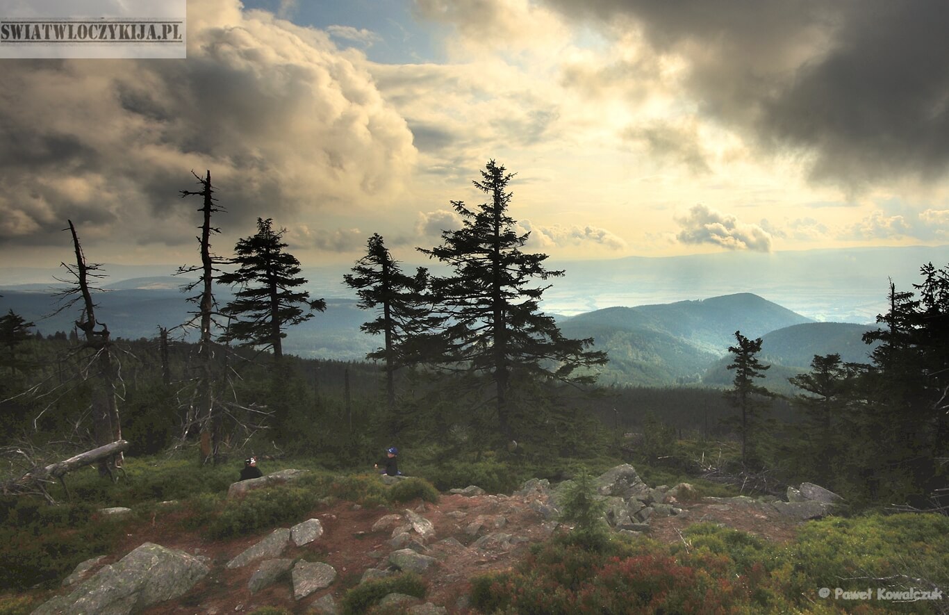 Obrazek przedstawia widok z Czarnej Góry. Na zdjęciu skalista ścieżka. Na drugim planie drzewa. Na trzecim planie góry i zachmurzone niebo z którego rozbłyska światło